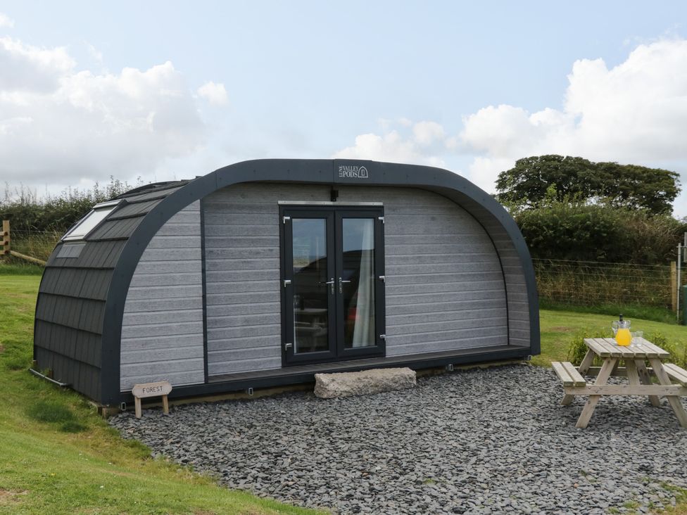 A curved wooden pod with double glass doors on a gravel area next to a picnic table with a jug of juice at Forest in Dalton-In-Furness