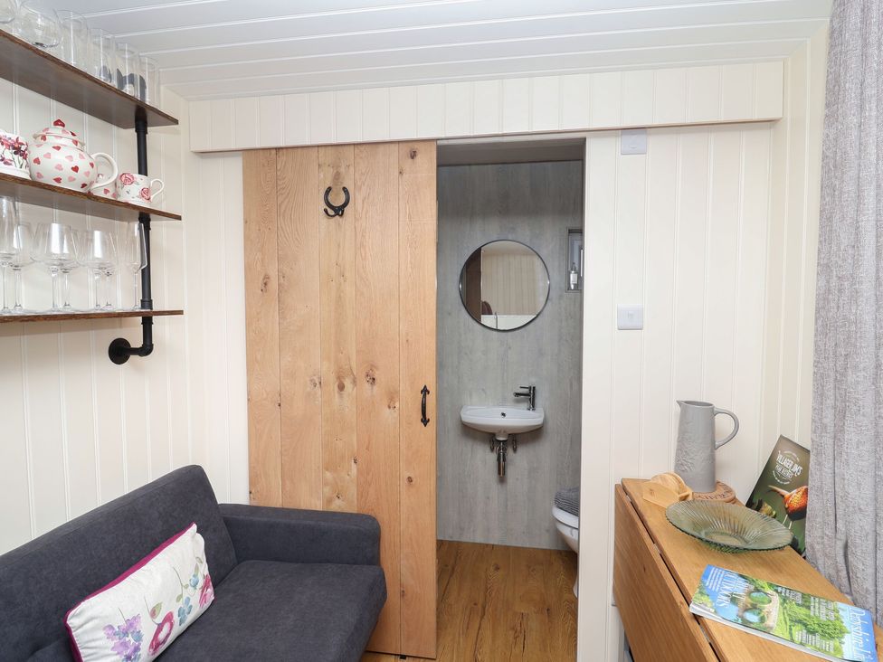 A small living area with a gray couch a sliding wood door leading to a bathroom with a round mirror and sink at Hillside Hut in Radbourne near Ashbourne
