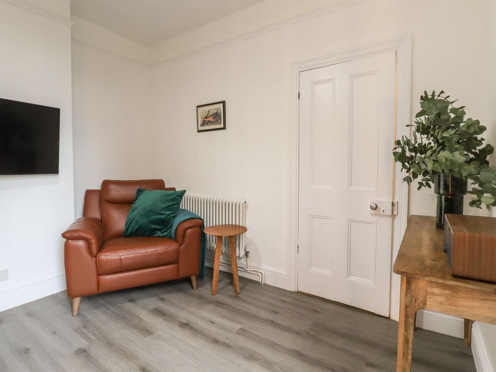 A corner of a living room with a brown armchair and green pillow a wall-mounted TV a wooden side table a plant on a wooden console table and a closed white door at Mabel in Whitstable