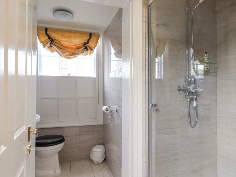 A bathroom with a toilet, a window with a yellow roman shade, and a glass-enclosed shower at 3 Old Market Place in Harleston
