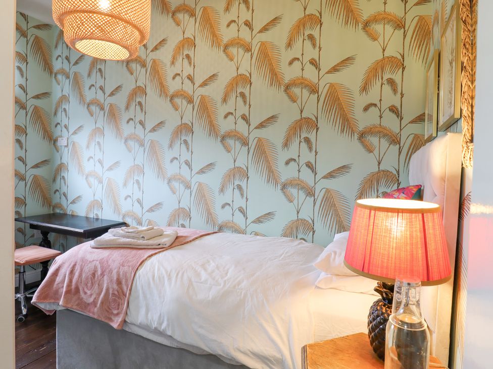 A bedroom with a single bed a table with a lamp and a glass bottle and palm leaf wallpaper at 3 Old Market Place in Harleston