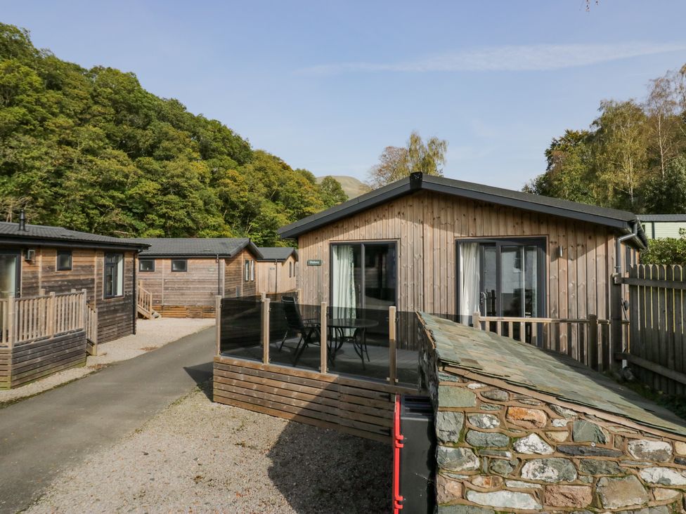 A group of wooden cabins with glass balconies and outdoor tables on a paved path with trees in the background at The Bothy in Keswick