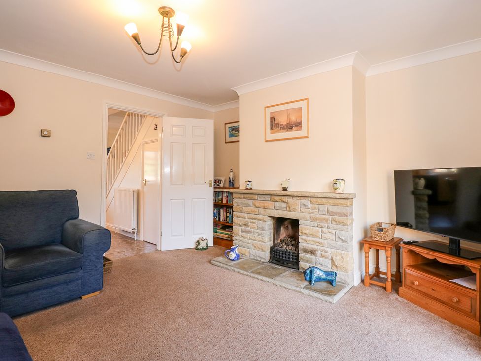 A living room with a fireplace, armchairs, a TV on a wooden stand, and an open door showing stairs at 1 Crowlands Cottages in Holt