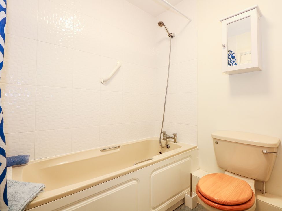 A bathroom with a beige bathtub with a showerhead above it a toilet with a wooden seat and a wall-mounted white cabinet with a mirror at 1 Crowlands Cottages in Holt