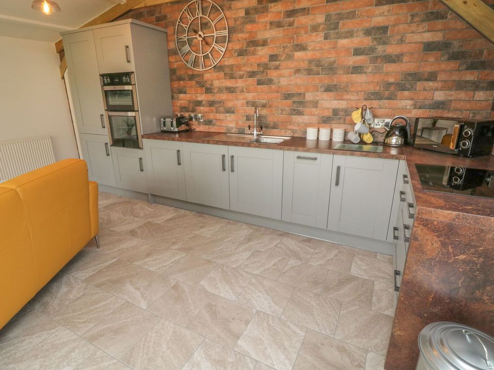 A kitchen with grey cabinets, brick wall, yellow sofa, built-in oven, sink, microwave, kettle, toaster, and tiled floor at Windy Willows in Okehampton