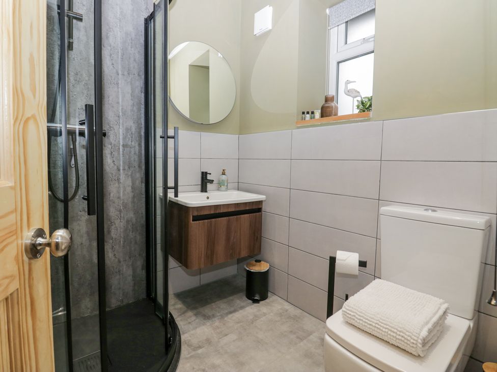A bathroom with a shower cubicle a wall-mounted sink a round mirror a toilet with folded towels and a window with toiletries on the sill at Willow Cottage in Silverdale