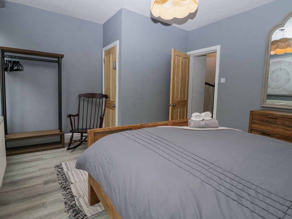 A bedroom with a bed covered in gray bedding wooden rocking chair open wardrobe and wooden dresser with a mirror at Willow Cottage in Silverdale
