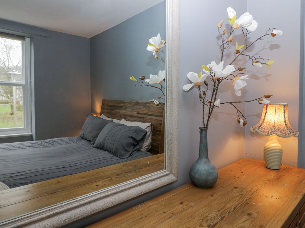 A bedroom with a wooden headboard bed reflected in a mirror and a wooden dresser with a vase of flowers and a lamp at Willow Cottage in Silverdale