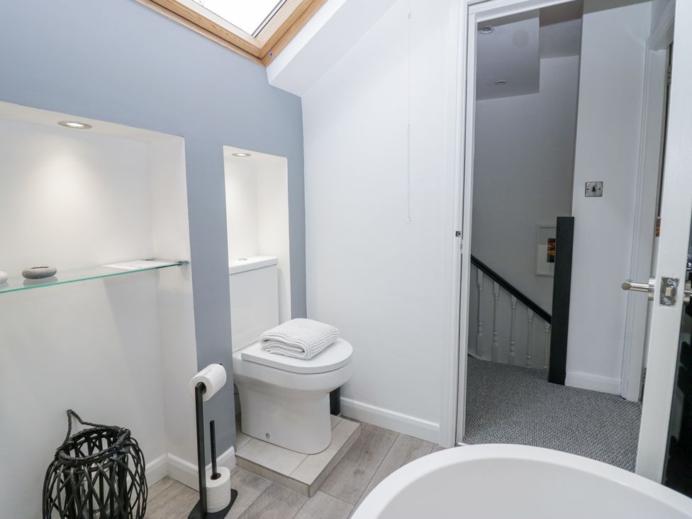A bathroom with a toilet, a folded towel on the toilet, a toilet paper holder, a black basket, a skylight window, and a doorway leading to a carpeted hallway at Willow Cottage in Silverdale