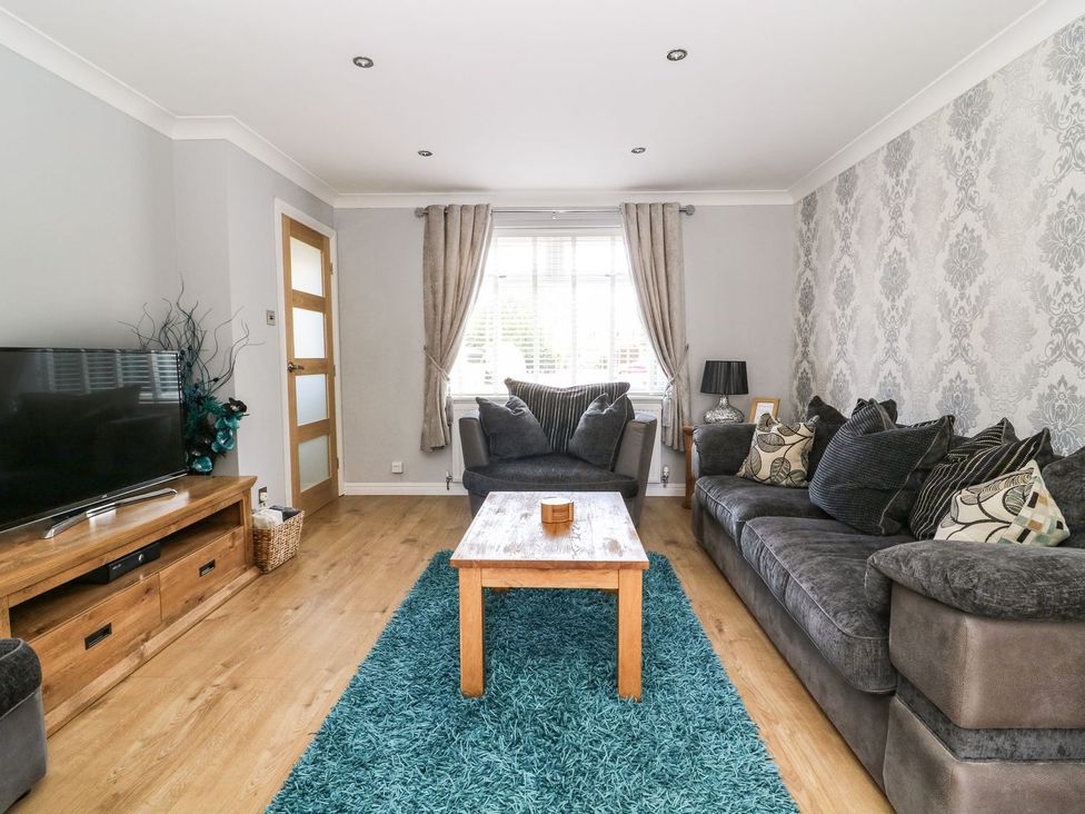 A living room with a large gray sofa and armchair a wooden coffee table a blue rug a wooden TV stand with a television and a window with curtains at The Whyte Retreat in Hornsea