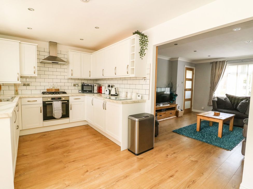 A kitchen with white cabinets and appliances connected to a living room with a wooden table on a blue rug at The Whyte Retreat in Hornsea