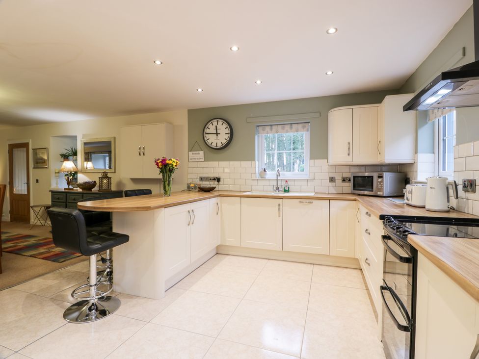 A kitchen with white cabinets wooden countertops a black bar stool flowers on the counter a clock on the wall and small appliances at Oakbank in Carrutherstown near Annan