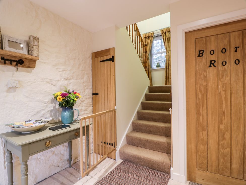 A hallway with stairs carpeted leading up with a window at the top a small table with flowers on it on the left and a door labeled boot room on the right at Oakbank in Carrutherstown near Annan