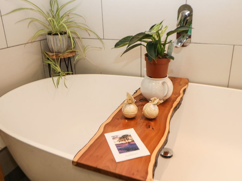 A bathtub with a wooden tray holding a small potted plant two wrapped bath bombs and a card with a picture of a tree in a field at Harebell Cottage in Curbar