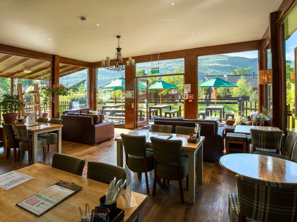 A dining area with tables, chairs, sofas, and large windows showing outdoor umbrellas and greenery at Ardgartan Argyll Golden Oak in Ardgartan, Loch Long