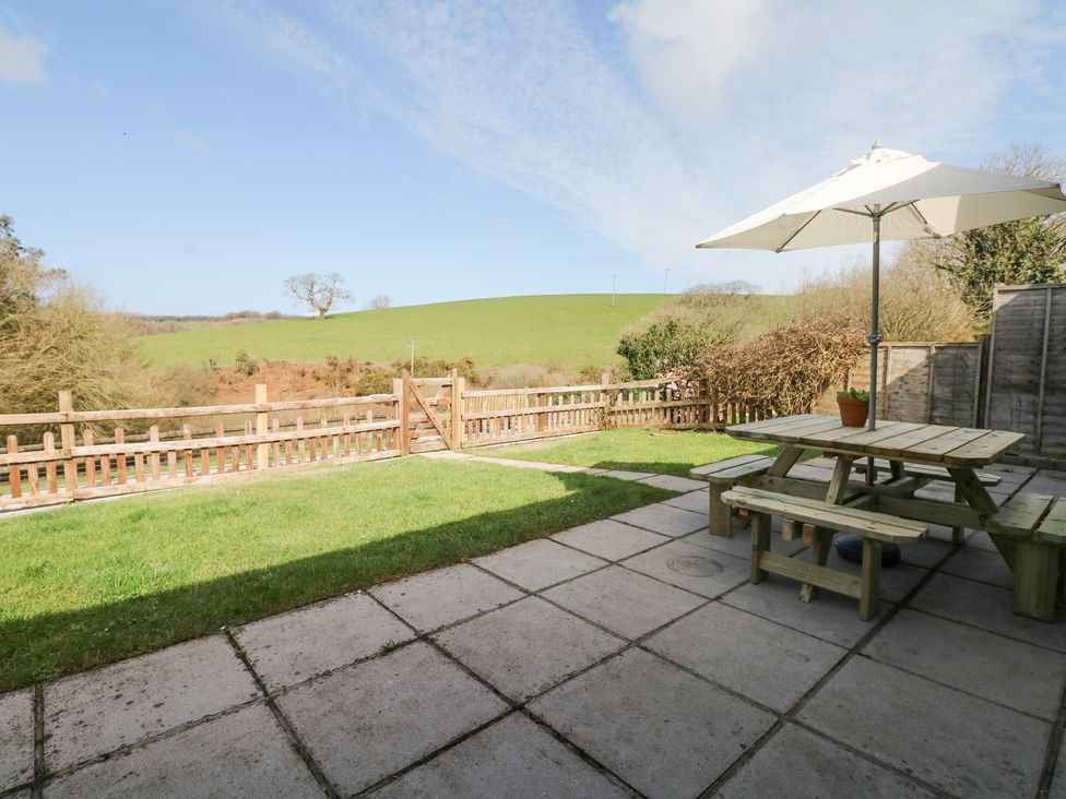 A patio with a wooden picnic table and umbrella next to a grass lawn and wooden fence with green hills in the background at Woodland View in Muddiford near West Down