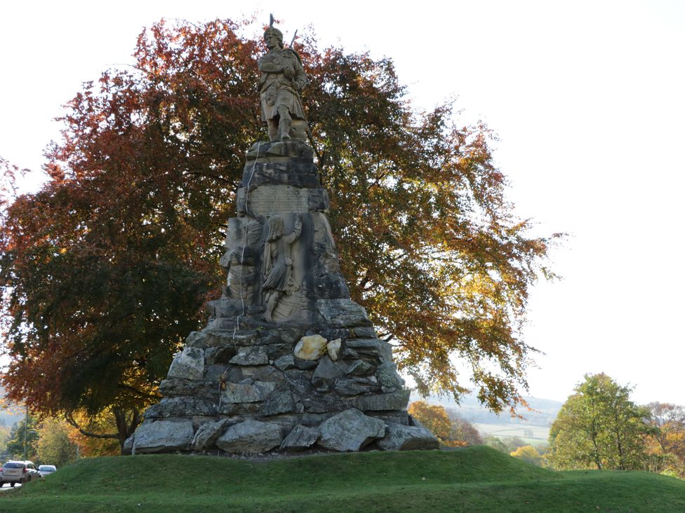 A stone statue monument with a figure on top and carved scenes at the base surrounded by trees and grass at 2 Strathtay Lodges in Aberfeldy