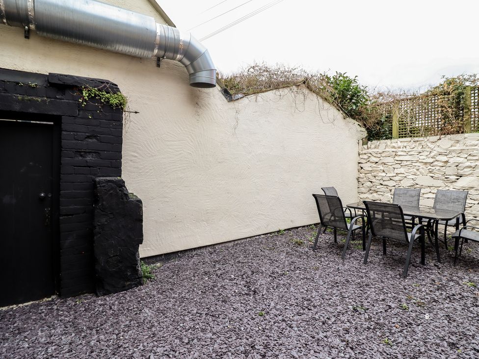 An outdoor patio area with a glass table and six chairs on a gravel ground with cream and stone walls at Gwydyr Bach 1 in Llanrwst