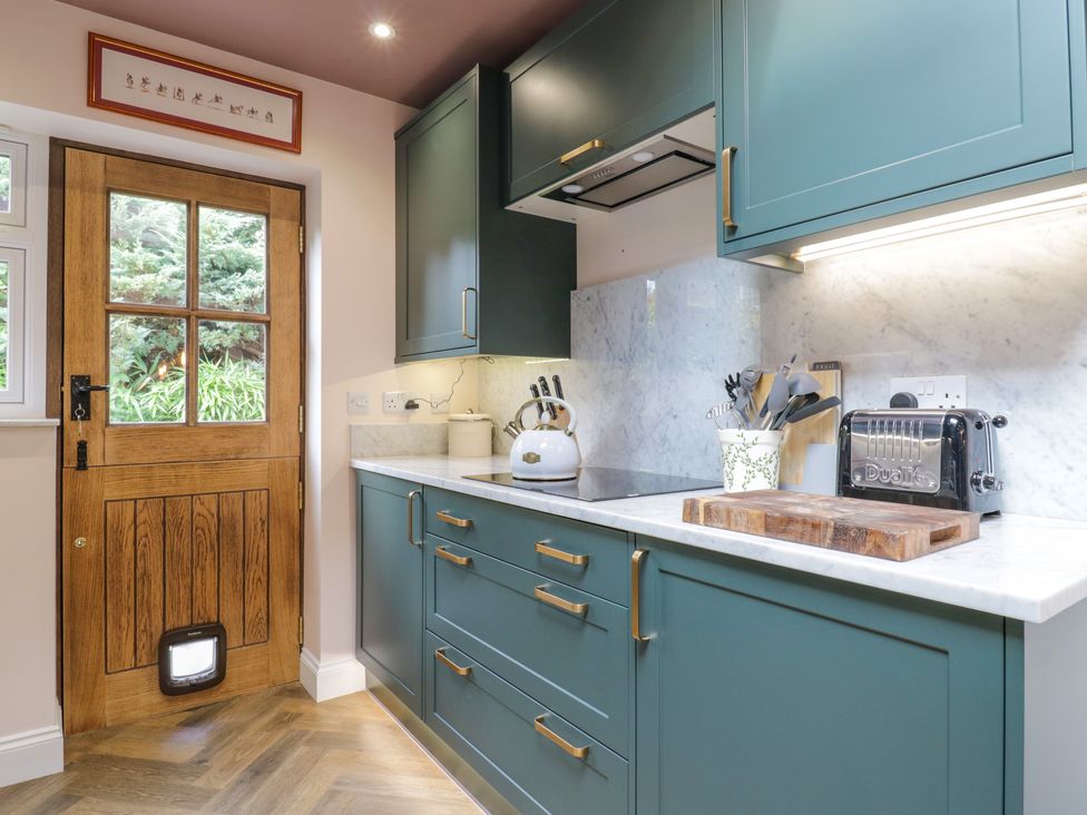 A kitchen with teal cabinets wooden door with glass panels and cat flap white kettle wooden cutting board and black toaster at Two Copthorne Cottages in Croxley Green