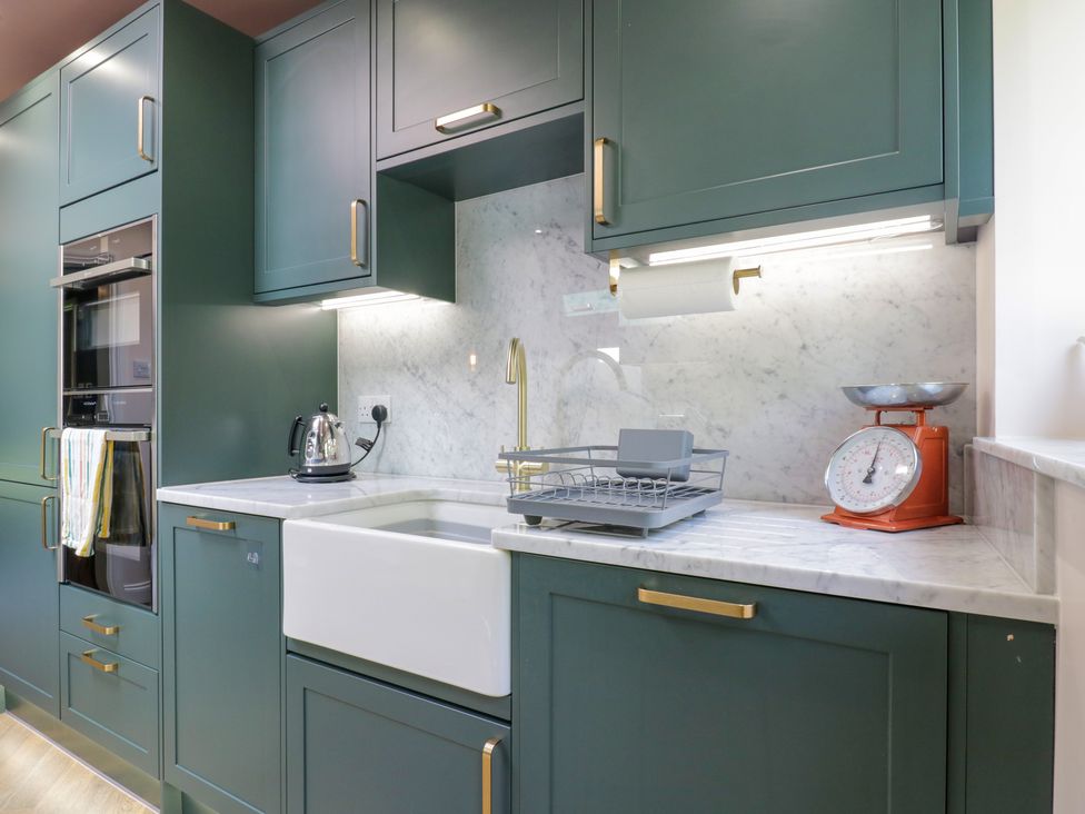 A kitchen with green cabinets a white sink a drying rack a kettle and an orange kitchen scale at Two Copthorne Cottages in Croxley Green