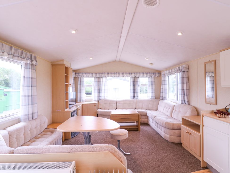 A living room with beige sofas a wooden table a TV on wooden shelves and windows with curtains at The Richmond in St Osyth