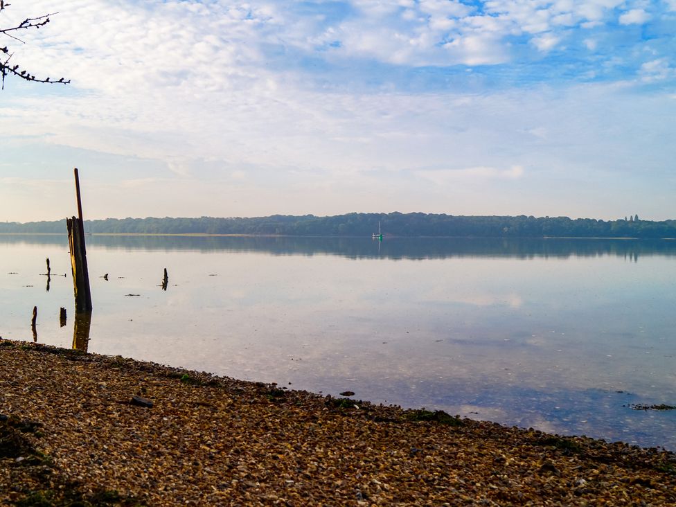 A calm lake with a pebble shore and wooden posts in the water with trees in the distance