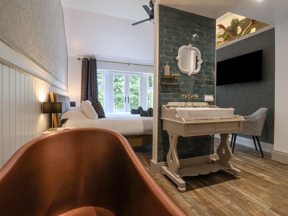A bedroom with a copper bathtub a white washstand a bed with pillows and bedside lamps a wall-mounted television and wooden flooring at Waters Edge Lock View in Gargrave