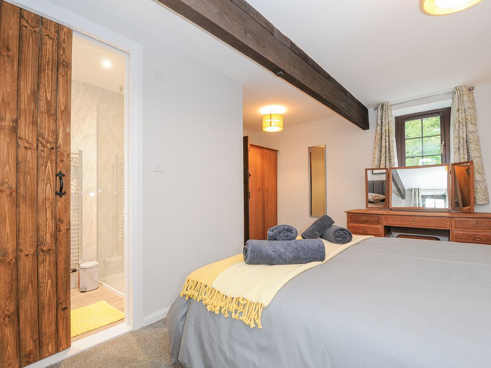 A bedroom with grey bedding and towels on the bed a wooden dressing table with mirrors a window wooden framed door leading to a bathroom with a yellow rug at Manor Barn in Saltash