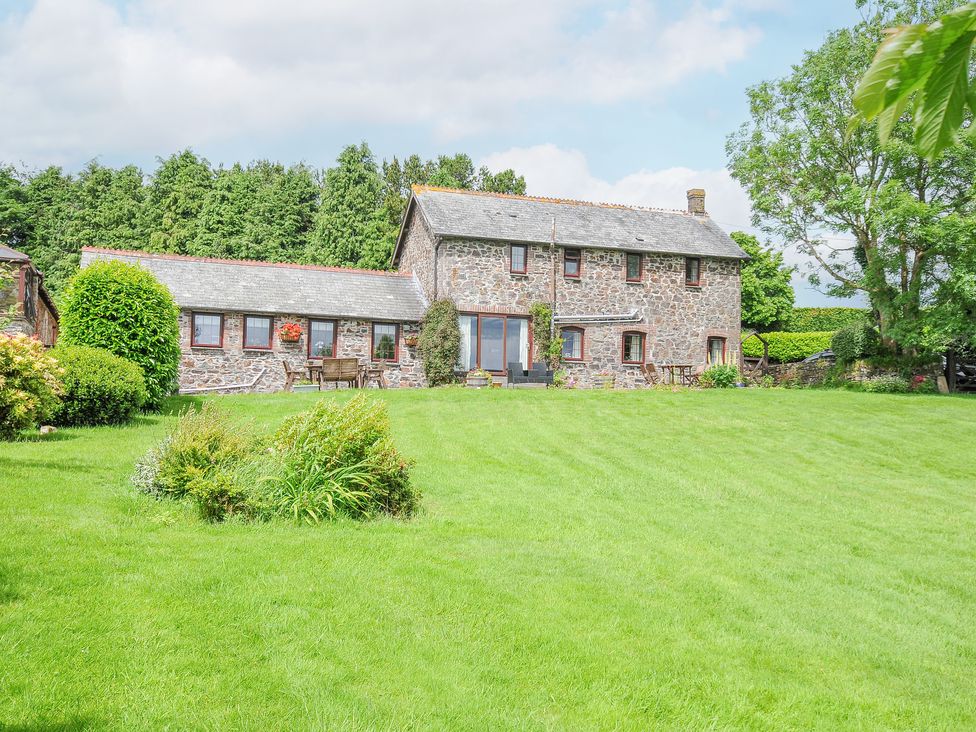 A stone house with a large green lawn and outdoor furniture in front surrounded by trees and bushes at Manor Barn in Saltash