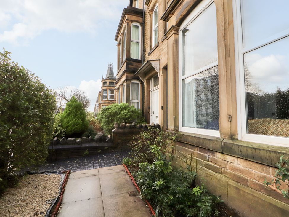 An exterior view of a stone building with windows and a front door alongside a paved walkway with bushes and small trees at Inglemead in Morecambe