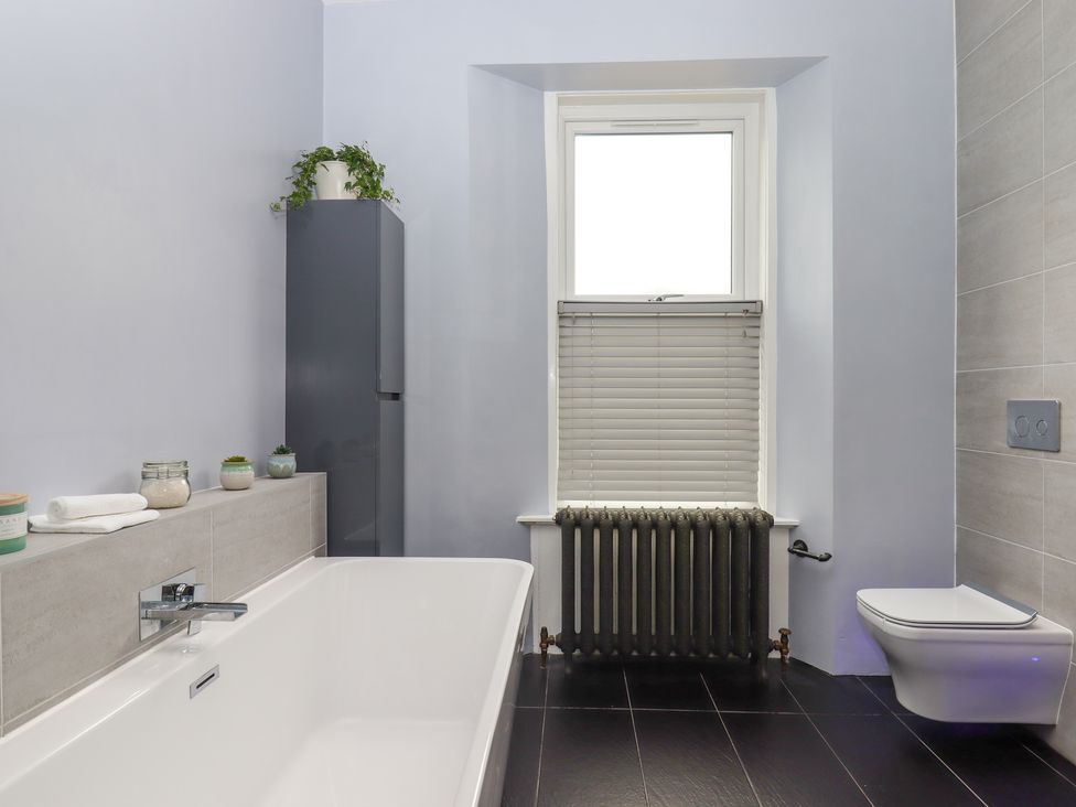 A bathroom with a bathtub, a tall cabinet with a plant, a window with blinds, a radiator, and a wall-mounted toilet at Rockland House in Brixham