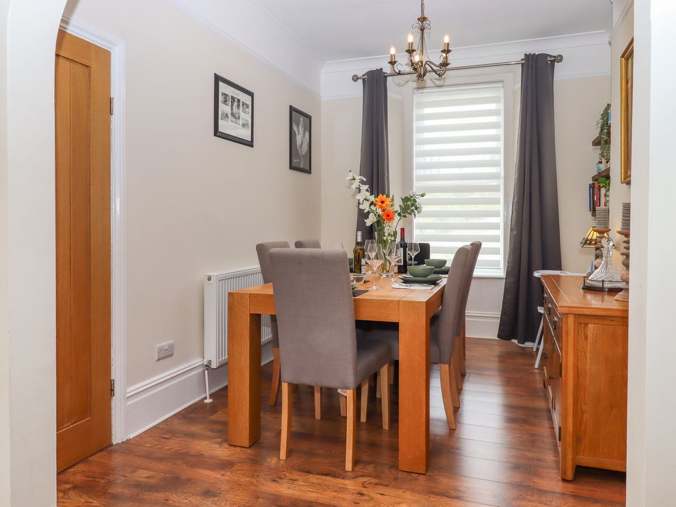 A dining room with a wooden table set with dishes and glasses six chairs wooden sideboard and window with curtains at Rockland House in Brixham