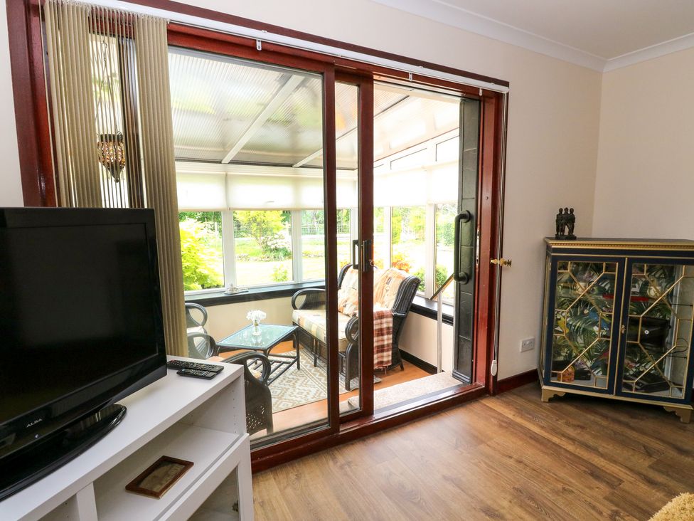 A living room with a television and glass cabinet next to sliding doors leading to a sunroom with wicker furniture at Mo Dhachaidh in Roy Bridge