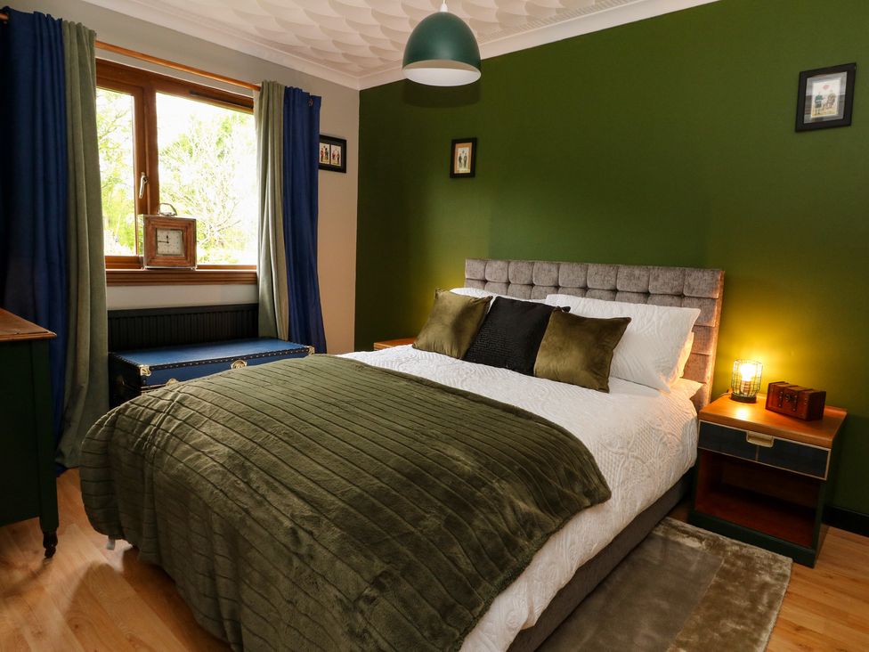 A bedroom with a bed a bedside table a window with curtains and framed pictures on the wall at Mo Dhachaidh in Roy Bridge