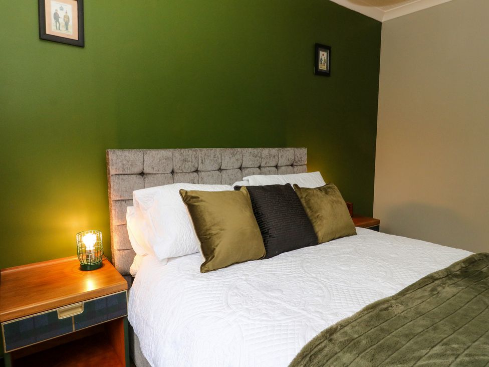 A bedroom with a bed covered in white bedding with green and black pillows beside wooden nightstands with lamps on a green wall at Mo Dhachaidh in Roy Bridge