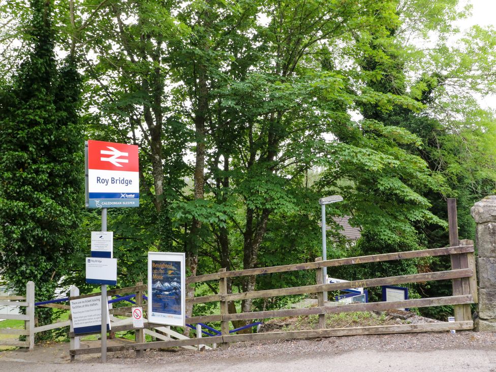 A railway station entrance with signs and a wooden fence surrounded by trees at Mo Dhachaidh in Roy Bridge