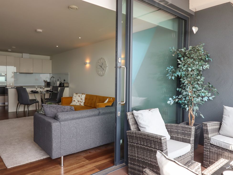 A living area with gray and orange sofas next to a dining table and kitchen with white cabinets and an outdoor seating area with wicker chairs and a potted plant at Apartment 17 Abbey Sands in Torquay