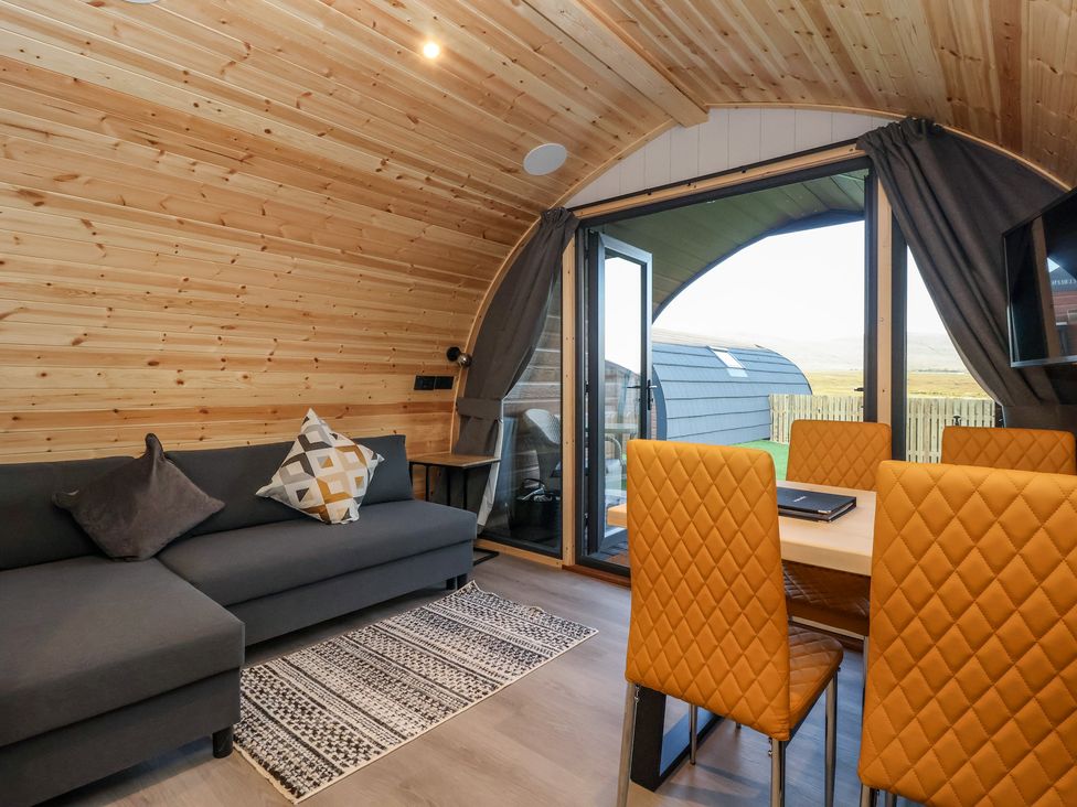 A wooden interior living space with a dark grey sofa and patterned cushions near orange quilted dining chairs around a table at Barn Owl in Chapel Le Dale near Ingleton