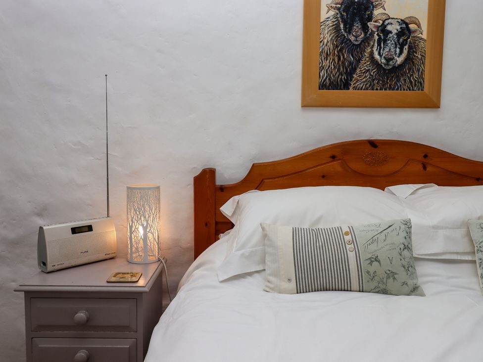 A bedroom with a wooden headboard bed white bedding two patterned pillows a bedside table with a lamp and a radio and a painting of two sheep on the wall at Primrose Cottage in Drewsteignton