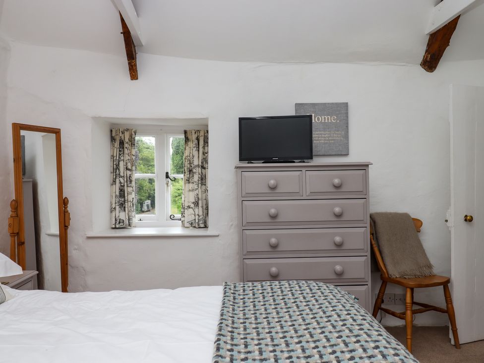 A bedroom with a bed a window with curtains a chest of drawers a chair with a blanket and a mirror at Primrose Cottage in Drewsteignton