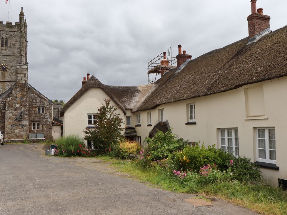 A thatched roof cottage with garden flowers and a stone church with clock tower in the background at Primrose Cottage in Drewsteignton