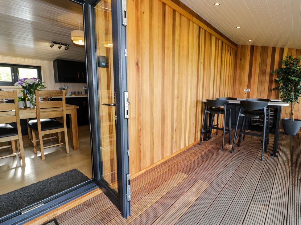 A wooden patio with a table and black chairs next to a dining room with wooden chairs and a table at Enia Lodge 2 in Llanelidan near Corwen