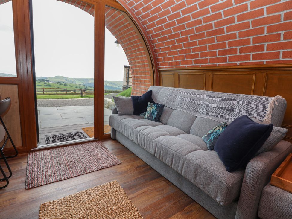 A living area with a gray sofa placed next to a large arched glass door showing outdoor hills and wooden fence at Rhosydd in Cyffylliog near Ruthin
