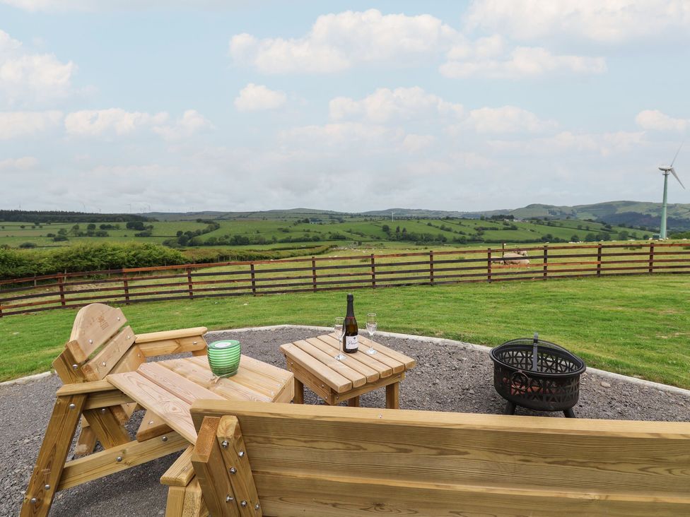 An outdoor seating area with wooden benches a table a bottle and glasses next to a fire pit with a view of green fields and hills at Rhosydd in Cyffylliog near Ruthin