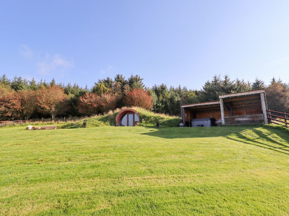 A grass-covered hobbit-style house door and a wooden shelter on a grassy hill near trees at Cae Bach in Cyffylliog near Ruthin