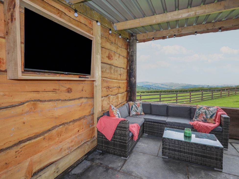 An outdoor covered seating area with a mounted television sofa with cushions and throw blankets and a glass coffee table overlooking a fenced green field at Weirglodd in Ruthin