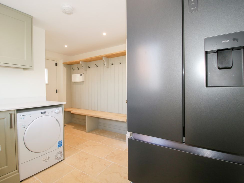 A utility room with a dryer beneath a countertop a black refrigerator and a wooden bench with wall hooks at Daniels Farmhouse in Claines
