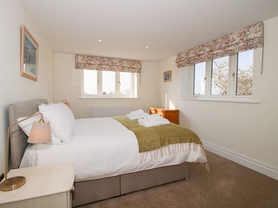 A bedroom with a bed covered with white and green bedding two windows with floral curtains bedside table and lamp a wooden chest of drawers on carpeted floor at Daniels Farmhouse in Claines