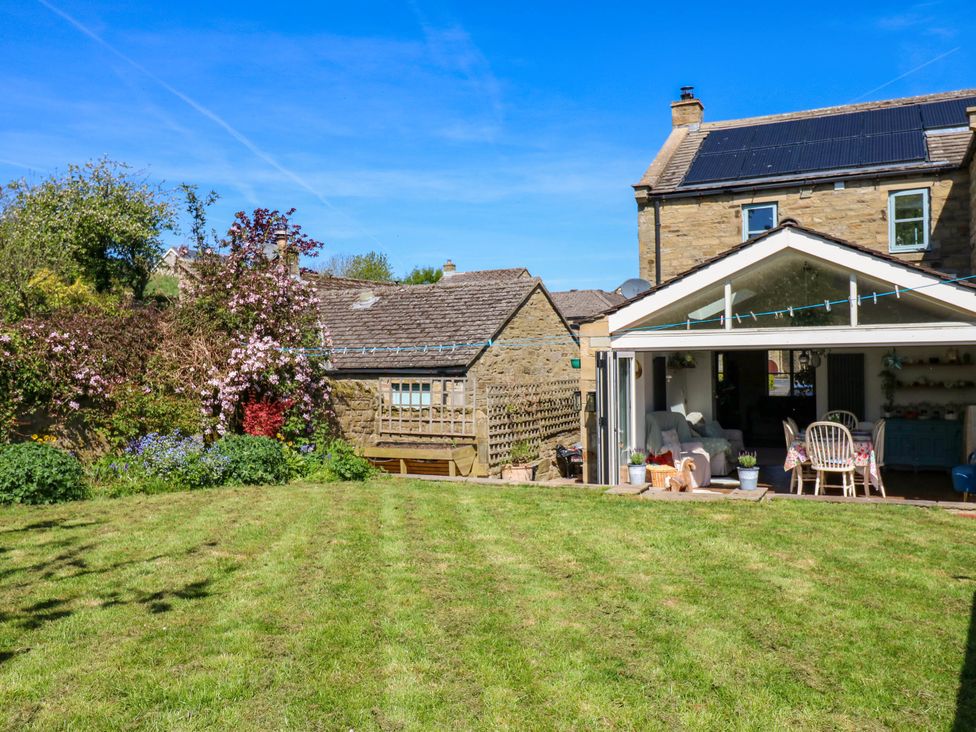 A garden with grass lawn and flowers next to a stone house with a glass extension and solar panels on the roof at 14 Hill Close in Reeth