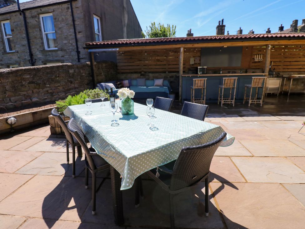 An outdoor patio with a dining table covered in a polka dot tablecloth surrounded by chairs and a bar area with stools and a seating area at 14 Hill Close in Reeth